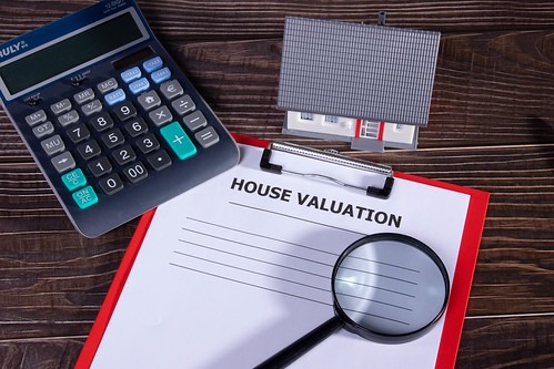 A paper with the words "House Valuation" in bold on a clipboard sitting on a desk surrounded by a calculator, magnifying glass and miniature house object