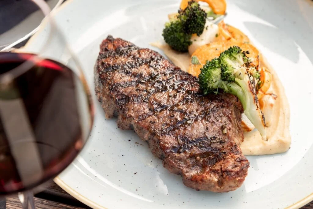 Steak with a side of potatoes sitting on a plate next to a glass of wine