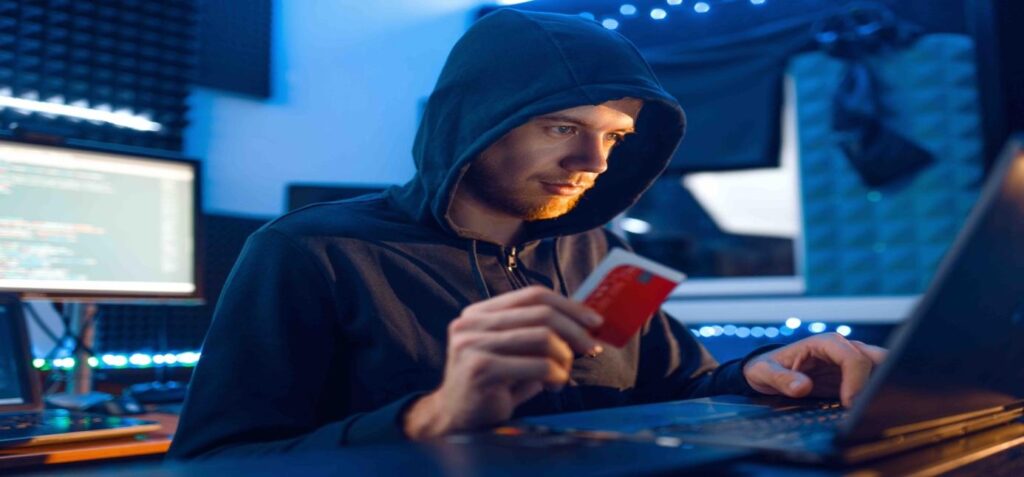a man at a desk in a black hoodie holding a credit card while using the computer
