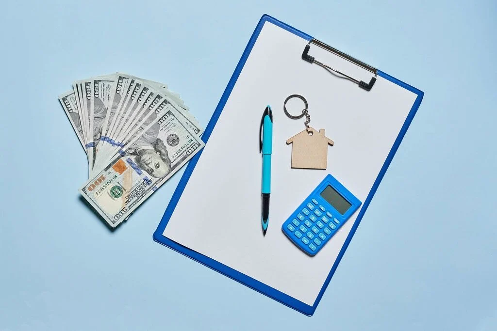stack of money sitting next to a clipboard that has a house key, pen, and calculator on top of it